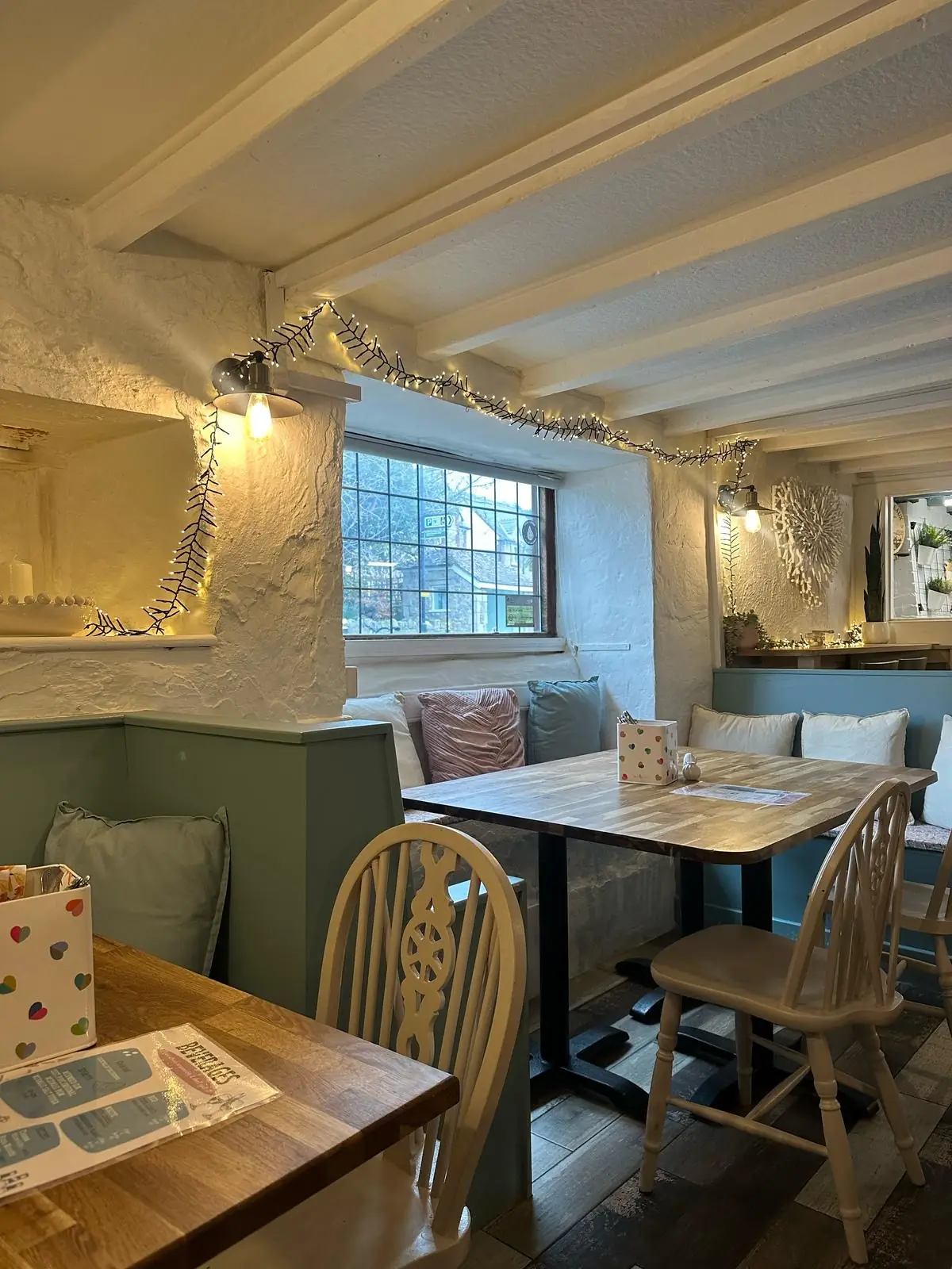 Cosy dining nook inside Calon with whitewashed stone walls, sage-green panelled benches, pastel cushions, fairy lights and a leaded cottage window